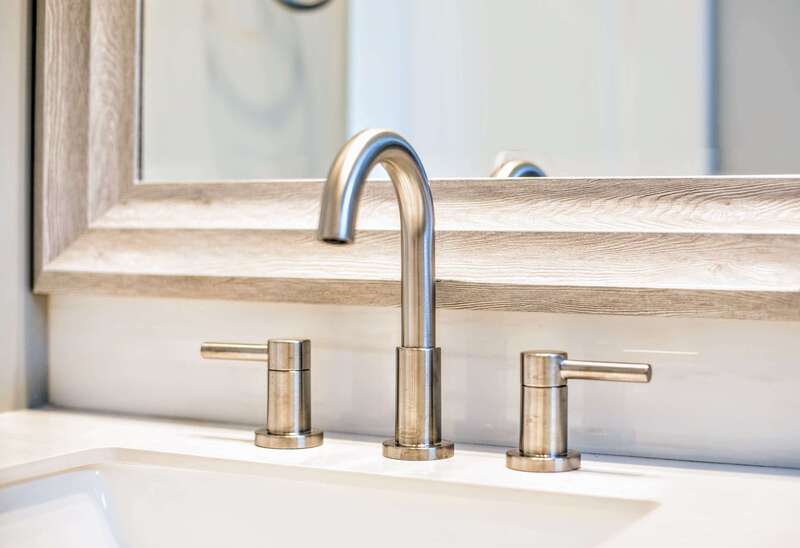 detailed high-end curved stainless steel bathroom faucet with pull handles, wood-framed mirror, and white counter and sink